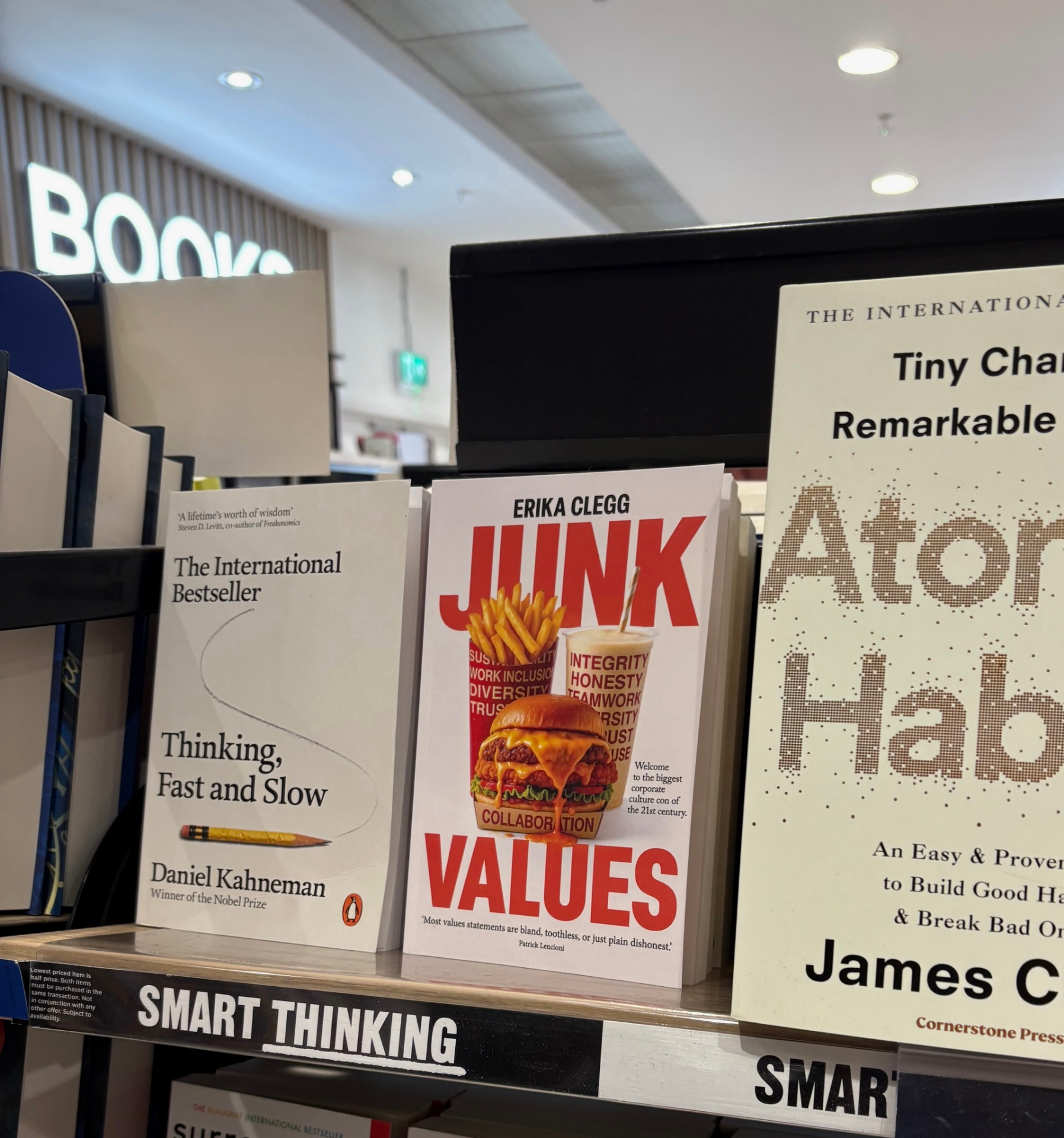Junk Values by Erika Clegg on display in a bookshop between Thinking, Fast and Slow and Atomic Habits